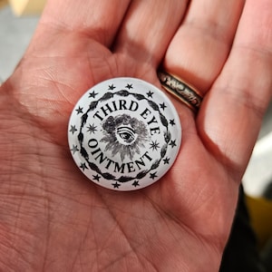 May include: A white button with a black and white design. The design features a stylised eye with the text "THIRD EYE OINTMENT" around it.