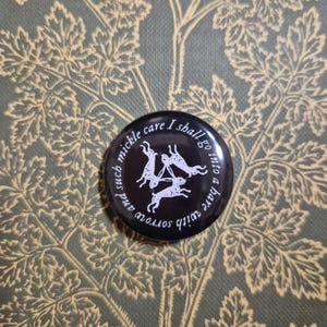 May include: A black button with white text and illustrations. The button features a circular design with the words "mickle care I shall go into a hare with sorrow and such" and illustrations of rabbits.