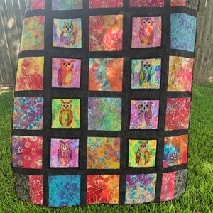 May include: A colorful quilt with a black background and a grid pattern. Each square features a different patterned fabric with an owl design in the center. The quilt is made with a variety of bright colors, including pink, blue, green, yellow, and orange.