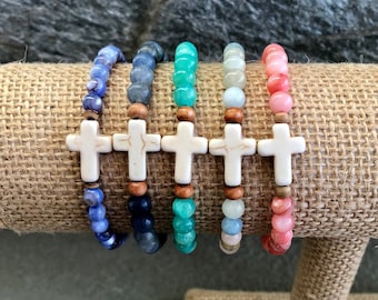 Op de afbeelding: Zes kralenarmbanden met een witte kruisbedel. De armbanden zijn in tinten blauw, groen en roze.
