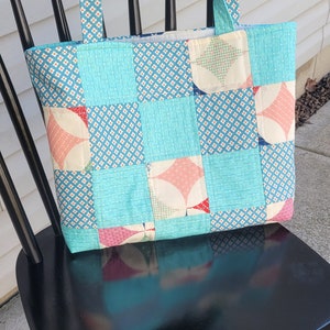 Pu&ograve; includere: Borsa tote fatta a mano con un design patchwork. La borsa presenta uno sfondo azzurro con vari motivi e forme nei toni del rosa, bianco e blu. I manici sono realizzati con lo stesso tessuto fantasia. La borsa &egrave; appoggiata su una sedia nera.
