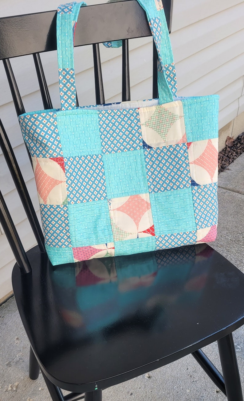 Pu&ograve; includere: Borsa tote fatta a mano con un design patchwork. La borsa presenta uno sfondo azzurro con vari motivi e forme nei toni del rosa, bianco e blu. I manici sono realizzati con lo stesso tessuto fantasia. La borsa &egrave; appoggiata su una sedia nera.