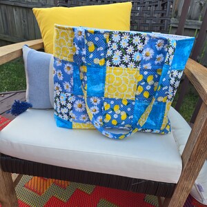 Pu&ograve; includere: Una borsa tote patchwork con un motivo floreale e a fette di limone blu, giallo e bianco. La borsa ha manici blu ed &egrave; appoggiata su un cuscino bianco. Un cuscino giallo e un cuscino a righe blu e bianche sono dietro.