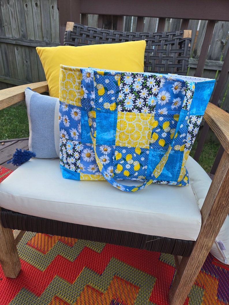 Pu&ograve; includere: Una borsa tote patchwork con un motivo floreale e a fette di limone blu, giallo e bianco. La borsa ha manici blu ed &egrave; appoggiata su un cuscino bianco. Un cuscino giallo e un cuscino a righe blu e bianche sono dietro.