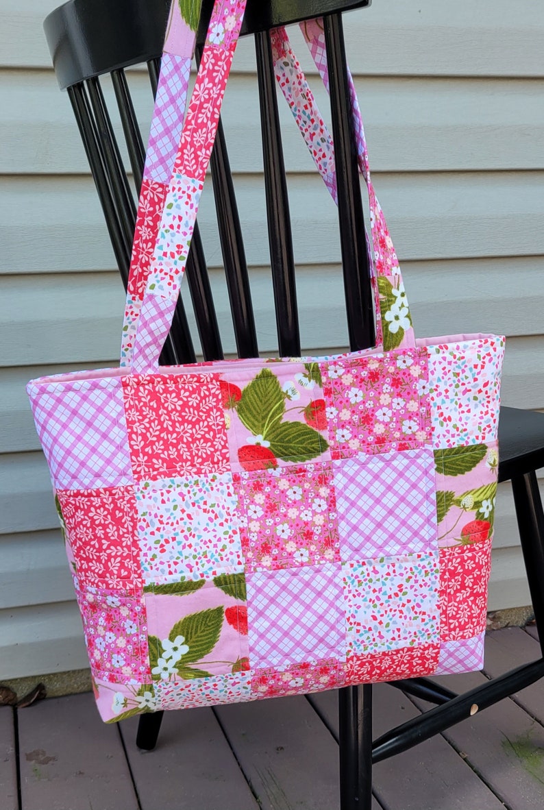 Pu&ograve; includere: Una borsa tote patchwork a tema fragola. La borsa presenta una variet&agrave; di tessuti rosa con motivi floreali e a quadri e motivi a fragola. La borsa ha cinghie abbinate ed &egrave; appoggiata su una sedia nera.