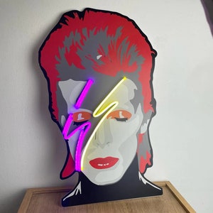Puede incluir: Un letrero de ne&oacute;n que representa la cara de David Bowie con un rayo en el ojo. El letrero est&aacute; iluminado con luces de ne&oacute;n rosa y amarillo.