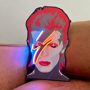 Puede incluir: Un letrero de ne&oacute;n que representa a David Bowie con un rayo en la cara. El letrero est&aacute; iluminado con luces de ne&oacute;n azules y amarillas.