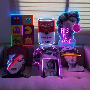Puede incluir: Una colecci&oacute;n de letreros de ne&oacute;n con varios dise&ntilde;os, que incluyen una lata de sopa Campbell's, un juego de Pac-Man, una cereza, un busto de David y un retrato de David Bowie. Los letreros est&aacute;n iluminados en varios colores, incluidos el rosa, el azul, el rojo y el amarillo.