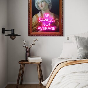 Puede incluir: Una obra de arte enmarcada con un retrato cl&aacute;sico y un letrero de ne&oacute;n. El letrero de ne&oacute;n rosa dice "BE SAVAGE, NOT AVERAGE". El marco adornado rodea la imagen. La obra de arte se exhibe sobre un taburete de madera y una cama.