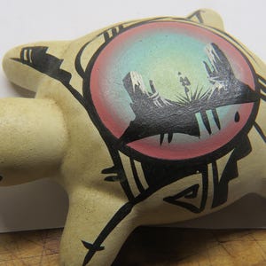 May include: A ceramic turtle figurine in a light beige color. The turtle's shell features a circular design with a desert landscape scene in shades of red, blue, and black. Black tribal-style lines and shapes decorate the shell.