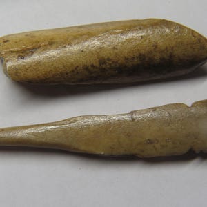 May include: Two antique bone tools. One is a rounded, oblong shape, and the other is a pointed awl with a notched handle. Both have a light tan color with darker areas.