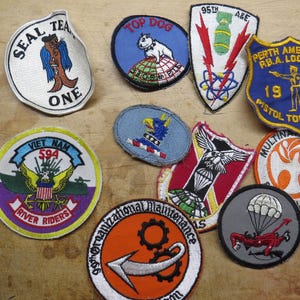 May include: A collection of embroidered patches with various designs and text. Patches include a seal with the text "SEAL TEA ONE", a dog with "TOP DOG", and a shield with "PERTH AMBOY, N.J. R.B.A. LOCAL 13 1968 PISTOL TOURNAMENT".
