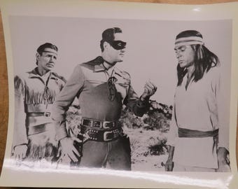 Op de afbeelding: Zwart-witfoto van drie mannen in een westernsetting. Eén man draagt een masker en een kostuum met een cape. De andere twee mannen zijn gekleed in westernkleding. De afbeelding is een vintage print.