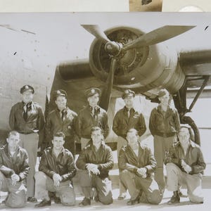 May include: Black and white photograph of a group of men in uniform posing in front of an airplane. The men are wearing jackets, caps, and ties. The airplane has a large propeller.