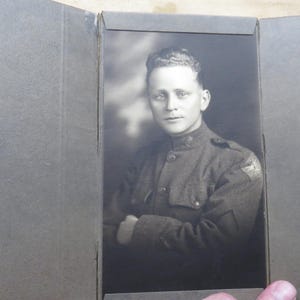 May include: A vintage black and white portrait of a man in a military uniform, likely from the early 20th century. The photograph is in a dark gray photo album. The man has short hair and is looking at the camera with his arms crossed.