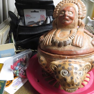 May include: A detailed, hand-carved ceramic box with a lid featuring a stylized human figure and a jaguar head. The box is brown with a red base. The image also shows a black bag, a pink plate, and other items.
