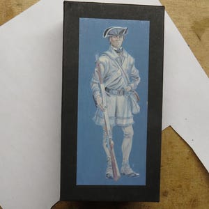 May include: A black rectangular box with a blue rectangular image of a colonial soldier. The soldier is wearing a white uniform, tricorn hat, and holding a rifle. The background is a light blue color.