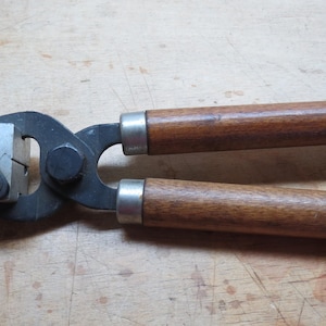 May include: A black metal bullet swaging tool with wooden handles. The tool has a lever and a metal block with a square opening.