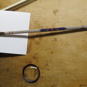 May include: An arrow with a wooden shaft, adorned with white beads, blue, red, and black accents. The fletching features brown feathers. The arrow is lying on a wooden surface.