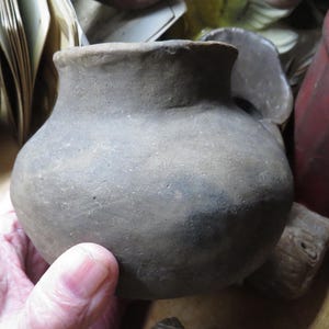 May include: A small, gray, earthenware pot with a rounded body and a slightly flared rim. The pot is held in a hand, and other pottery items are visible in the background, including a red container and a carved wooden figure.