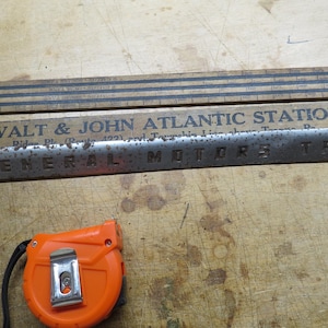 May include: An assortment of vintage tools and signage, including a wooden folding ruler with black stripes, a metal sign that reads "GENERAL MOTORS TRUCK", and an orange tape measure.