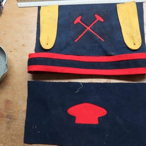 May include: A blue fabric hat with a red and black band. The hat has two yellow straps and a red embroidered emblem of two crossed pickaxes. The hat also has a red embroidered emblem of a red cap.