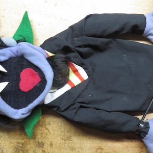 May include: A hand puppet featuring a black-haired character with a purple hood, white fangs, and a red heart-shaped mouth. The puppet wears a black jacket over a white and red striped shirt, with purple hands and green felt accents on the head.