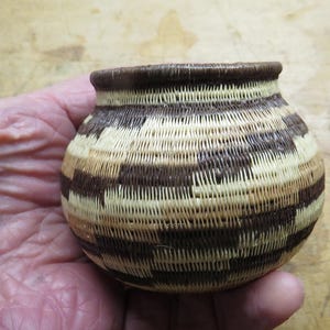 May include: A small, hand-woven basket with a rounded body and a slightly flared rim. The basket features a repeating pattern of alternating bands of light tan and dark brown fibers. The rim is a solid dark brown color.