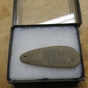 May include: A gray, teardrop-shaped stone carving with a floral design, displayed in a black box with a clear lid. The stone has a small hole at the top. The box is lined with white, fluffy material.