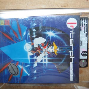 May include: A blue and red video game cartridge for the Amiga computer. The game is called "Interphase" and features a futuristic design with a woman in a red and blue outfit. The cartridge is in a clear plastic case.