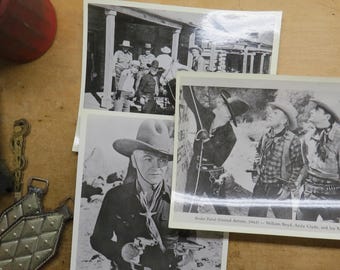 May include: Black and white photographs of cowboys. One photo shows a man in a cowboy hat holding a gun. Another shows a group of men in hats and cowboy attire. The photos are on a wooden surface.