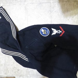 May include: A dark blue navy uniform jacket with white piping on the collar and a white star on the left side. The jacket has a patch on the right sleeve with the words "Seabees" and an eagle design.