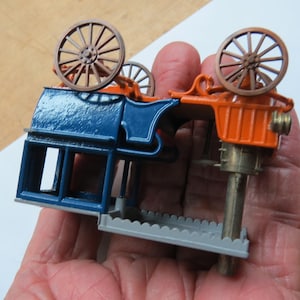 May include: A disassembled toy model of a vintage steam engine with blue, orange, and gray paint. The model features two large wheels and a detailed engine mechanism.