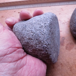 May include: A close-up of a gray, rough-textured stone with a rounded, slightly pointed top. The stone is being held in a hand.