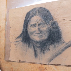 May include: A black and white pencil drawing of a person with long dark hair. The person is looking directly at the viewer with a serious expression. The drawing is on a piece of paper with a rough texture.