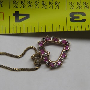 May include: Gold-toned necklace with a heart-shaped pendant. The pendant is adorned with pink gemstones and small white stones, creating a sparkling effect. The chain is delicate and the pendant is approximately 1 inch in size.