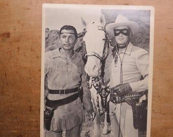 May include: Black and white photograph of two men in Western attire. One man wears a fringed shirt and belt with a holster. The other wears a mask, cowboy hat, and gloves, standing next to a white horse.