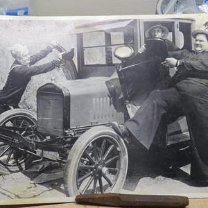 May include: Black and white photograph of three men interacting with a vintage car. One man is inside the car, while the other two are outside, appearing to struggle with the car's door. The car has large spoked wheels.