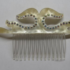 May include: Ivory-colored hair comb with a decorative top featuring a double-curve design. The comb is adorned with iridescent rhinestones and has a series of thin, clear teeth. A hair accessory.