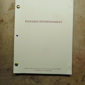 May include: A cream-colored document with the text "ENSEMBLE ENTERTAINMENT" in maroon. The document is held together with brass fasteners. The background is a wooden surface.
