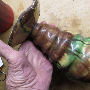 May include: A decorative, hand-turned wooden object with a wide, flat top and a bulbous, textured body. The object features a blend of brown, green, and yellow hues, with a glossy finish. The object is held in a hand.