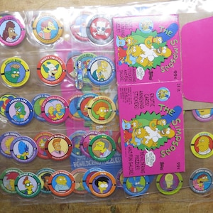 May include: A collection of The Simpsons trading cards and collectible discs. The discs feature various characters in colorful designs. Two pink boxes of stickers are also visible, with the text "The Simpsons" and "Exotic Cards and Fun Galore".