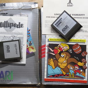 May include: Two Atari home computer game boxes featuring Millipede and Donkey Kong. Each set includes a black plastic game cartridge, a printed instruction manual, and a warranty card. The Donkey Kong manual displays a colorful illustration of the character.