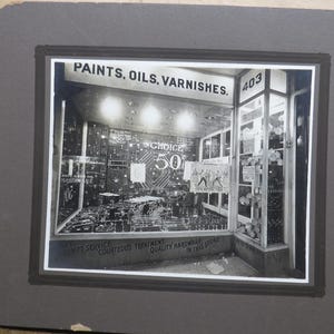 May include: Black and white photograph of a hardware store storefront. The sign reads "PAINTS, OILS, VARNISHES." A "Choice 50" sign is visible. The store displays various items, and the number "403" is on the window.