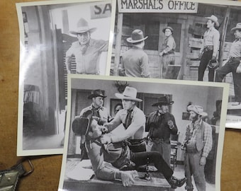 May include: A collection of vintage black and white photographs depicting scenes from a Western movie. The images feature actors in cowboy hats, vests, and shirts, with settings including a marshal's office and outdoor scenes.