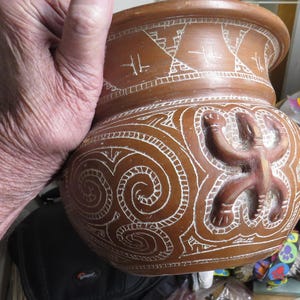 May include: A brown ceramic pot with white etched designs. The pot features a stylized human figure and spiral patterns. The pot is held by a person's hand. The pot has a wide opening and a rounded body.
