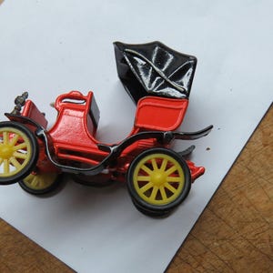 May include: A red toy car with a black fabric top. The car has yellow wheels and a detailed design.