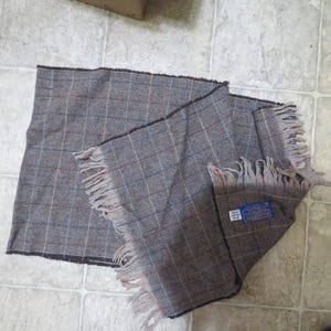 May include: A brown and grey plaid scarf with fringe on the edges. The scarf has a blue label with white text that reads "100% Pure Wool".