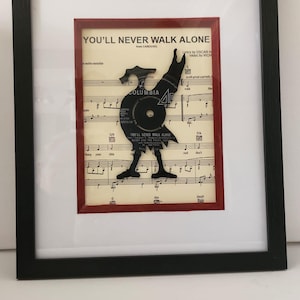 May include: A framed piece of art featuring a black vinyl record cut into the shape of a bird, standing on a sheet of music with the title "You'll Never Walk Alone" from the musical Carousel. The record is on a red background.
