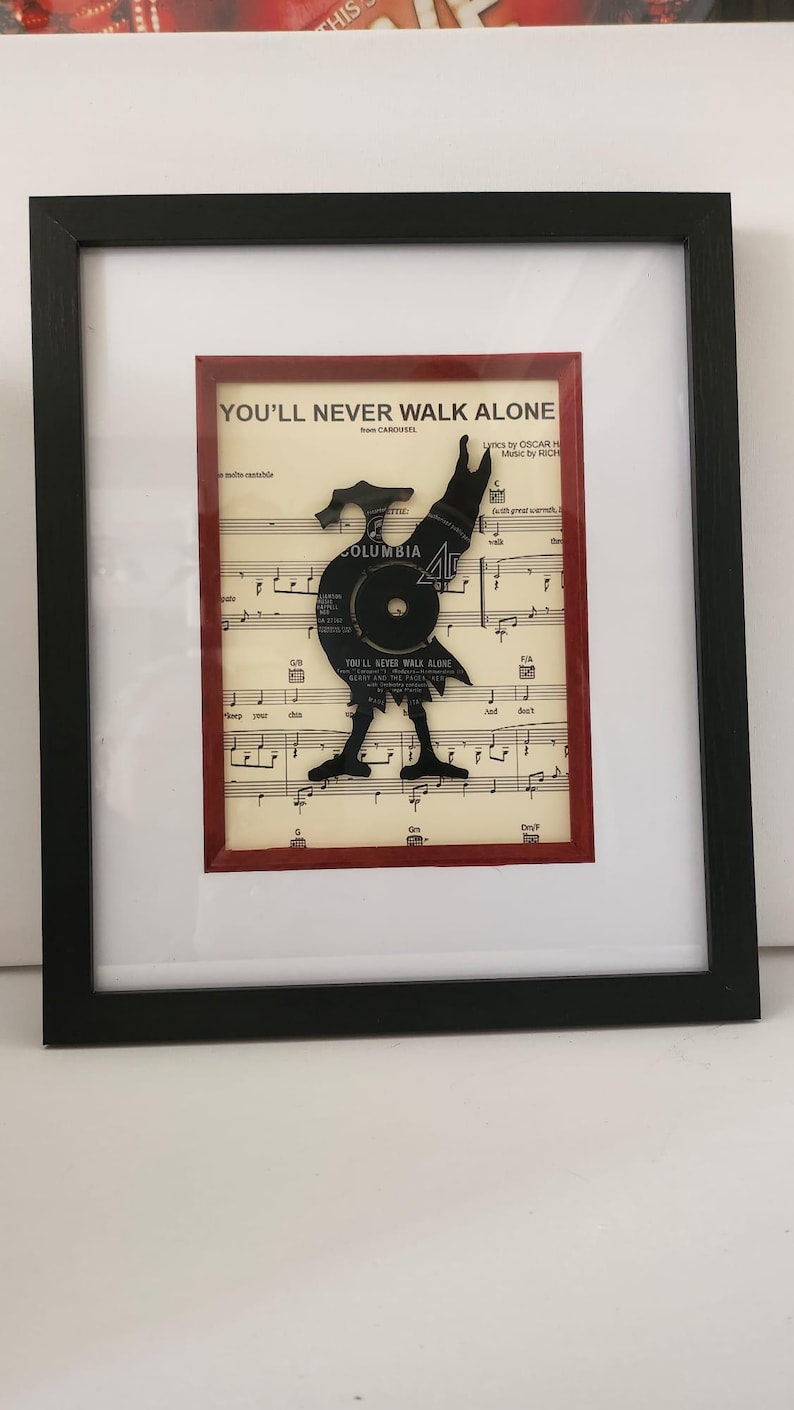 May include: A framed piece of art featuring a black vinyl record cut into the shape of a bird, standing on a sheet of music with the title "You'll Never Walk Alone" from the musical Carousel. The record is on a red background.